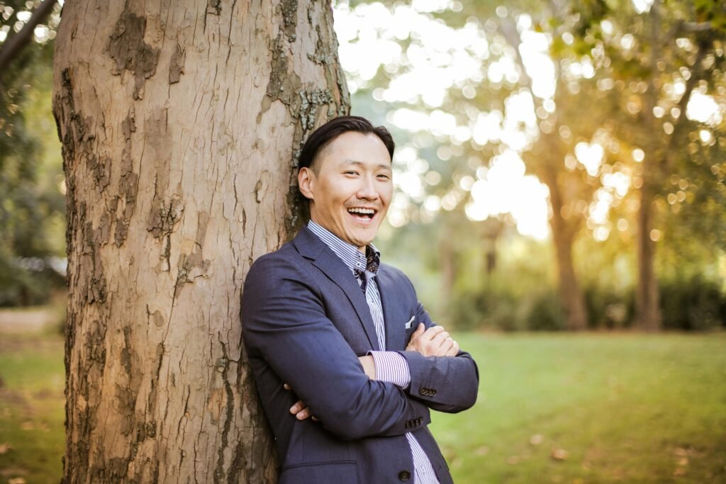 Smiling Asian man in a suit leaning against a tree in a park, enjoying the bright day.