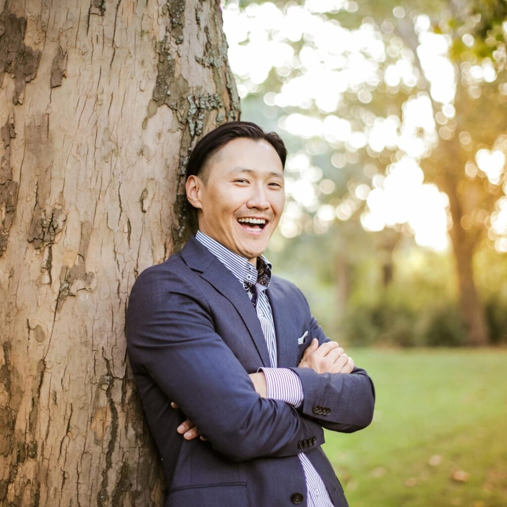 Smiling Asian man in a suit leaning against a tree in a park, enjoying the bright day.
