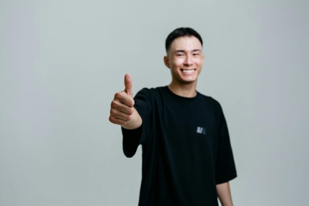 A cheerful young man in a black shirt smiles and gives a thumbs up in a studio setting.