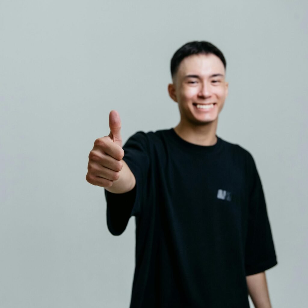 A cheerful young man in a black shirt smiles and gives a thumbs up in a studio setting.